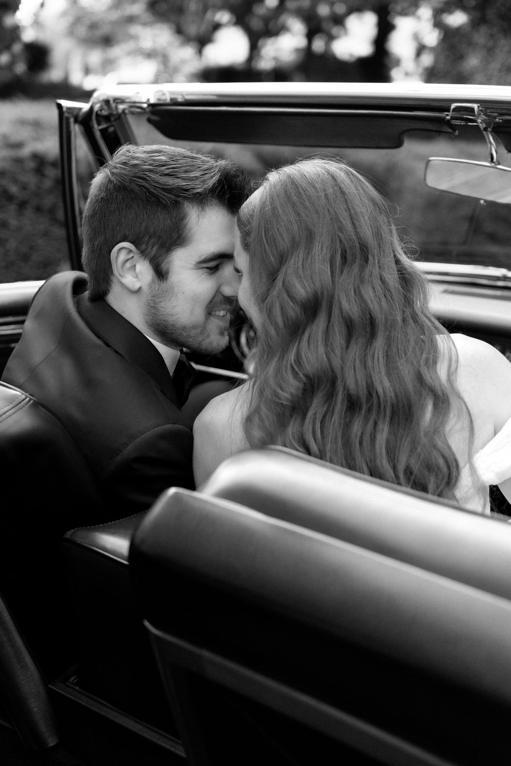 A romantic black and white photo of a couple sharing an intimate moment in a vintage convertible car.