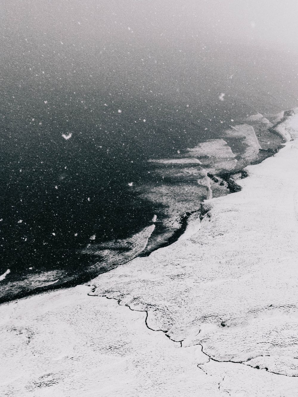 Aerial view of a snow-covered coastline with dark ocean waters and winding shoreline patterns visible from above.