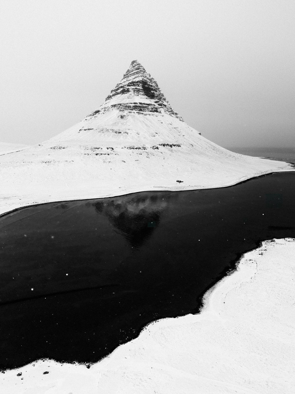 Black and white photo of snow-covered Kirkjufell mountain in Iceland with its reflection in a dark river below.