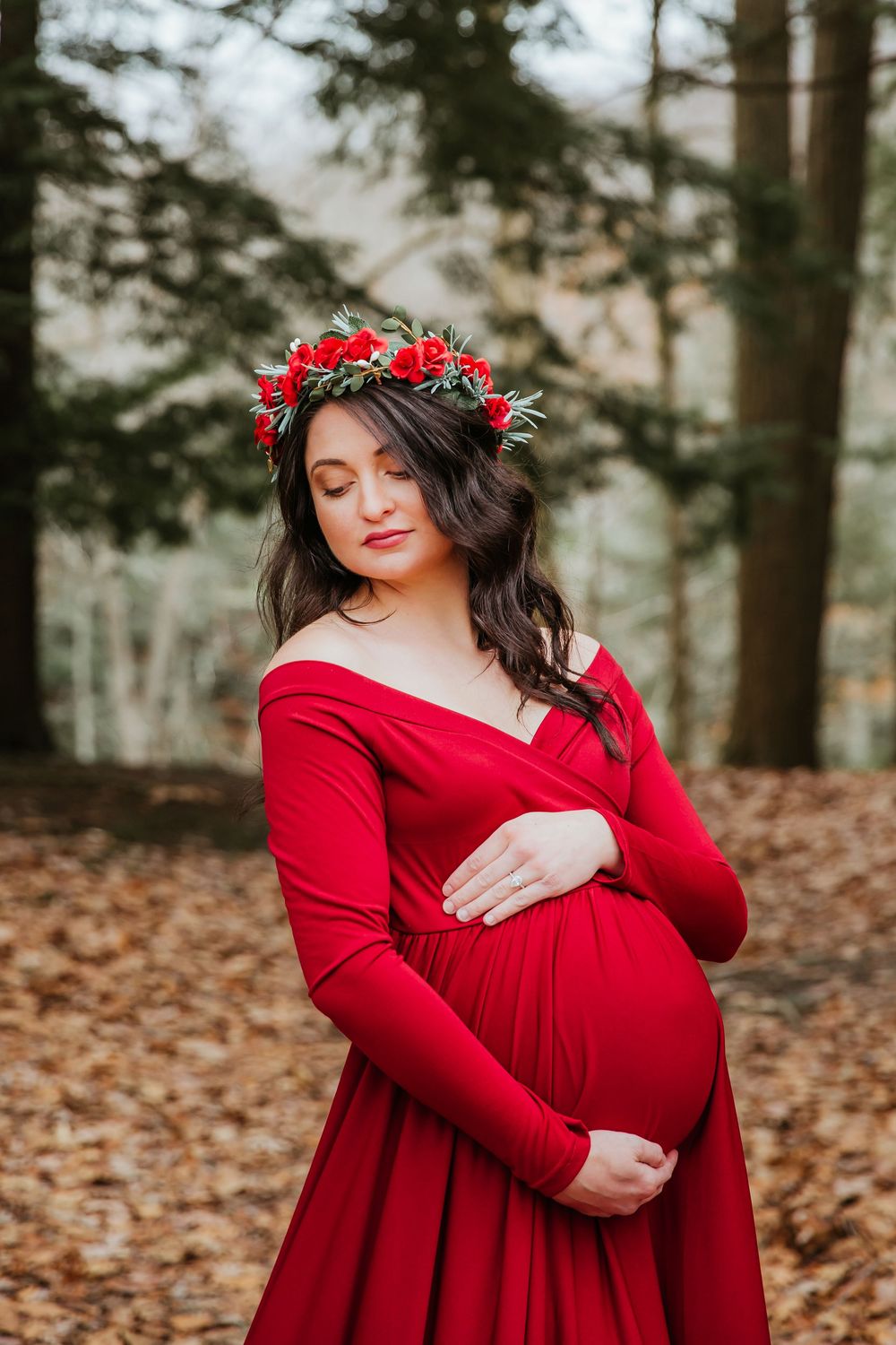Serene maternity portrait with red dress and floral crown in forest setting.