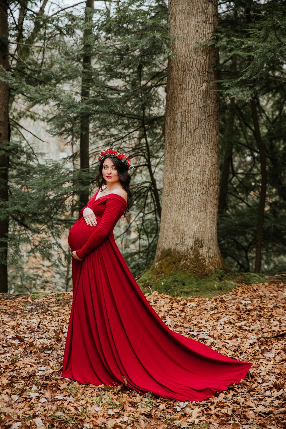 Dramatic maternity photo with flowing red dress train against evergreen forest.