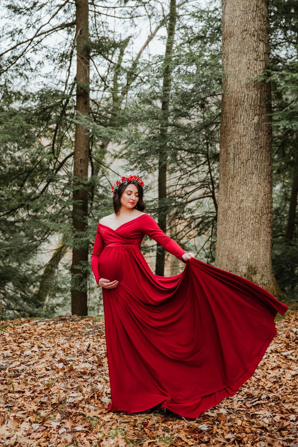 A flowing red maternity gown with off-shoulder design photographed in a misty forest setting with fallen leaves.