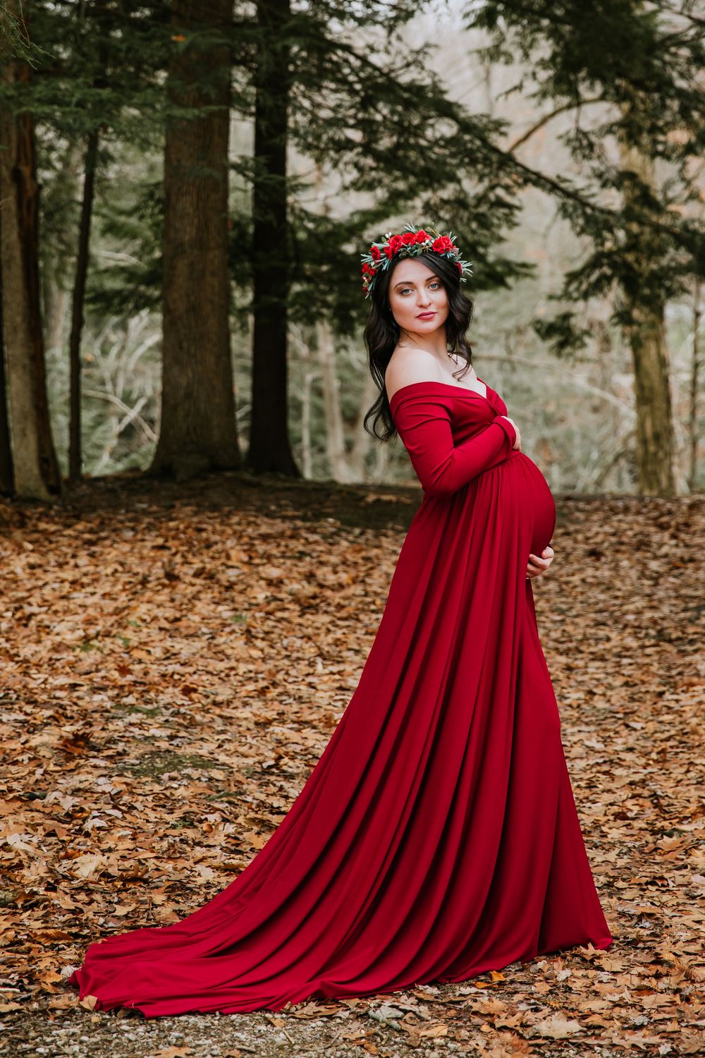Dramatic red maternity gown photographed among fall foliage and pine trees.