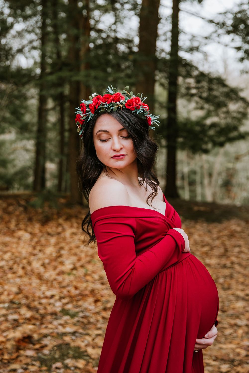 Maternity photo in forest featuring flowing red dress and floral crown against autumn leaves.
