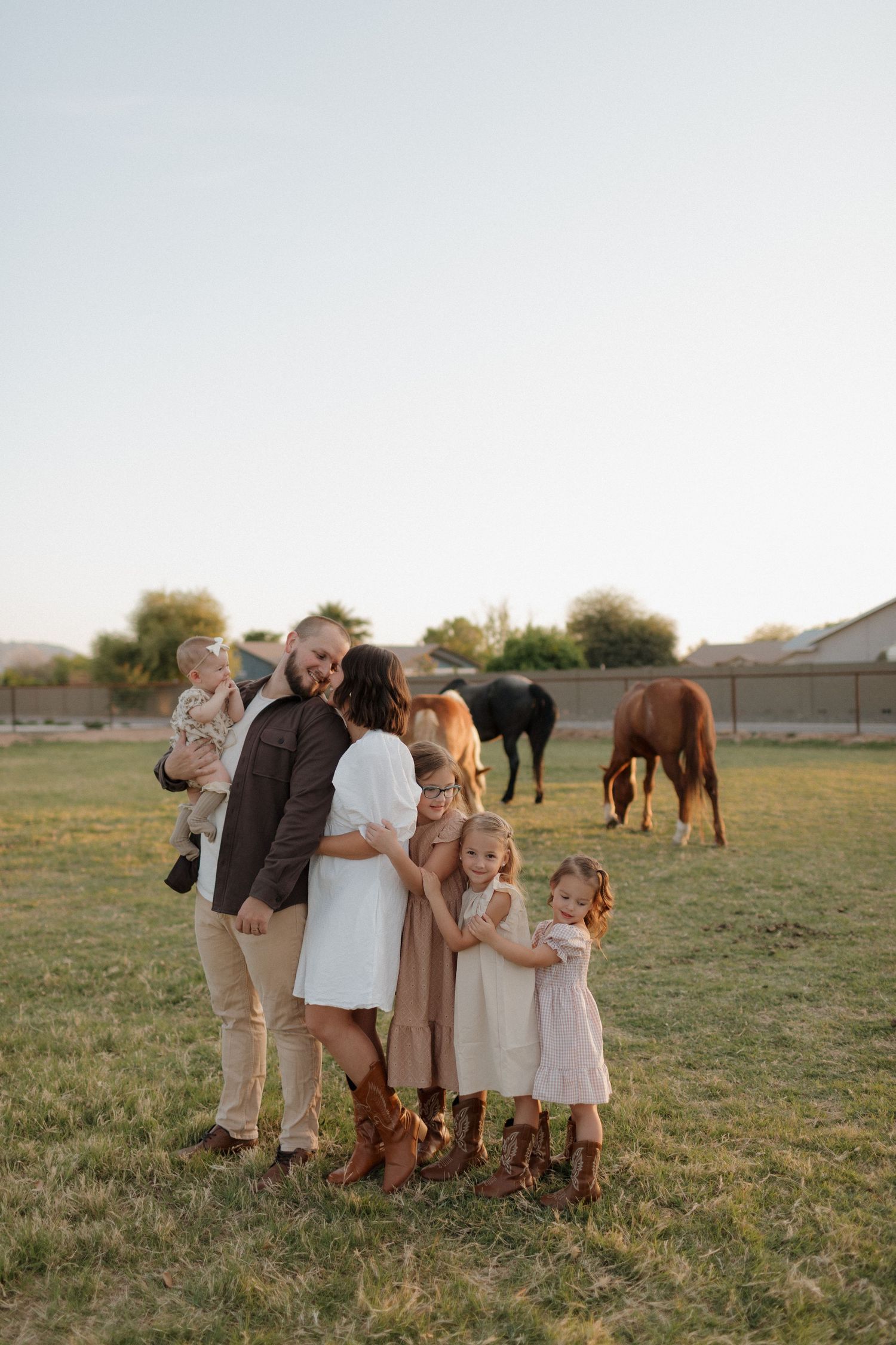 Family poses together in a field with horses grazing in the background at sunset.