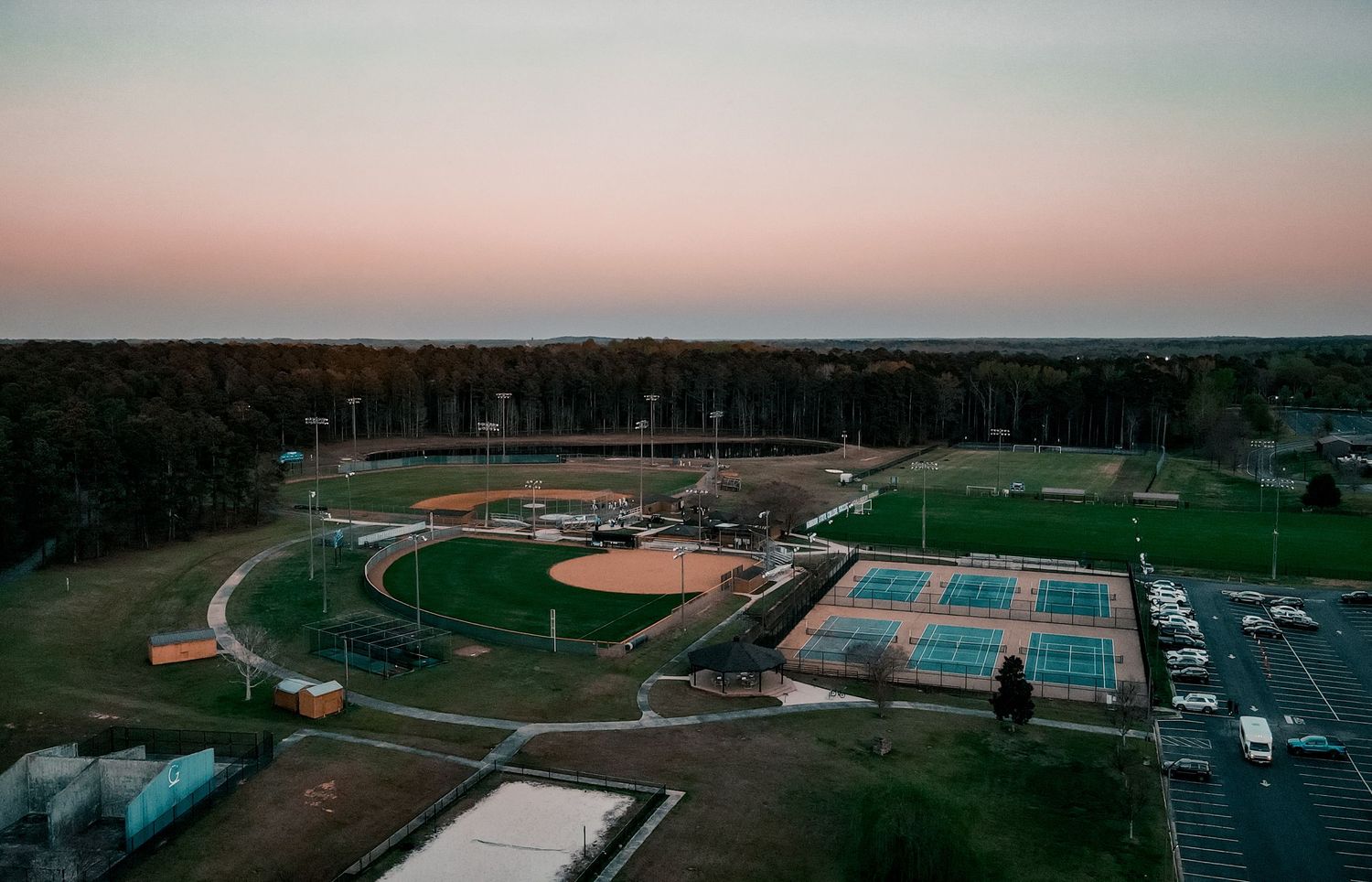 Aerial view of baseball diamond and tennis courts at dusk with soft pink sky above rural sports complex.