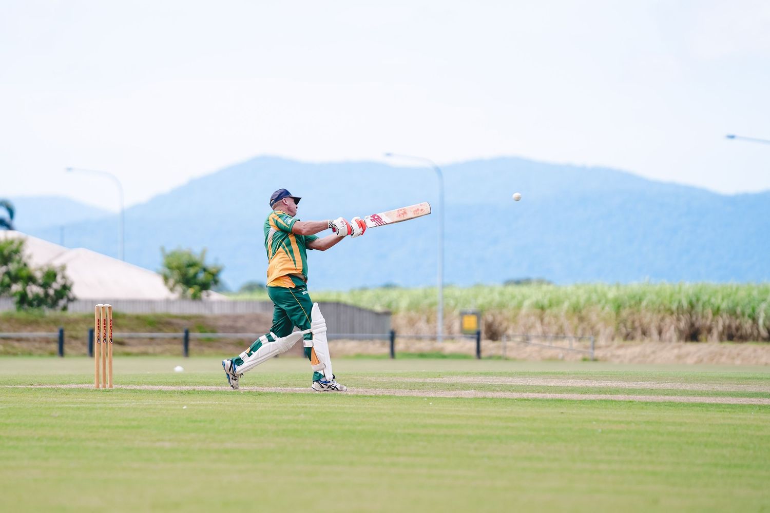 A cricket player runs in to bowl on a green grass field with mountains visible in the background.