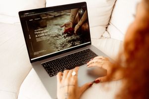 Hands typing on laptop keyboard showing website content on screen while sitting on white couch.