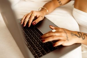 Tattooed hands typing on a laptop keyboard while wearing bright nail polish and delicate jewelry.