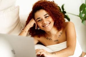 A person with curly red hair smiles while working on laptop in a bright indoor setting.