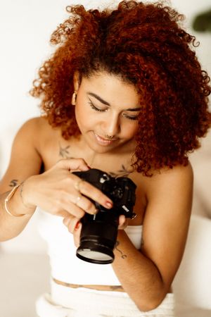 Photographer with vibrant red curly hair examining a digital camera while wearing a white top.