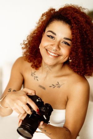 Professional photographer wearing a white top and displaying beautiful curly red hair holds a DSLR camera.