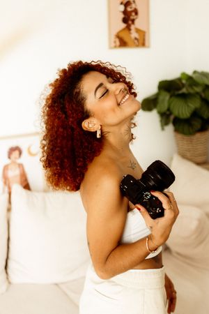 Creative photographer in white outfit poses joyfully with camera in a bright studio setting with decor.