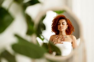 Reflection in mirror shows person with curly red hair in white top amid green plant leaves.