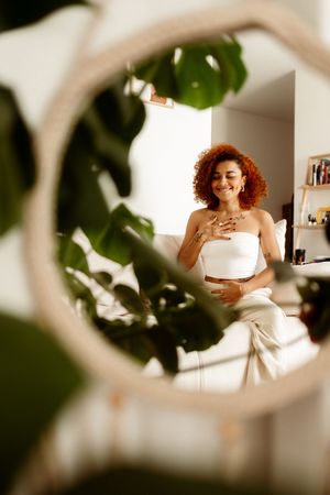 Through indoor plants, a mirror reflects someone in a white dress with curly red hair smiling.