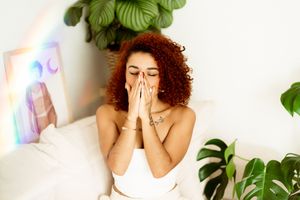 Person with curly red hair in white top reacting with surprise expression against white wall with tropical plants.