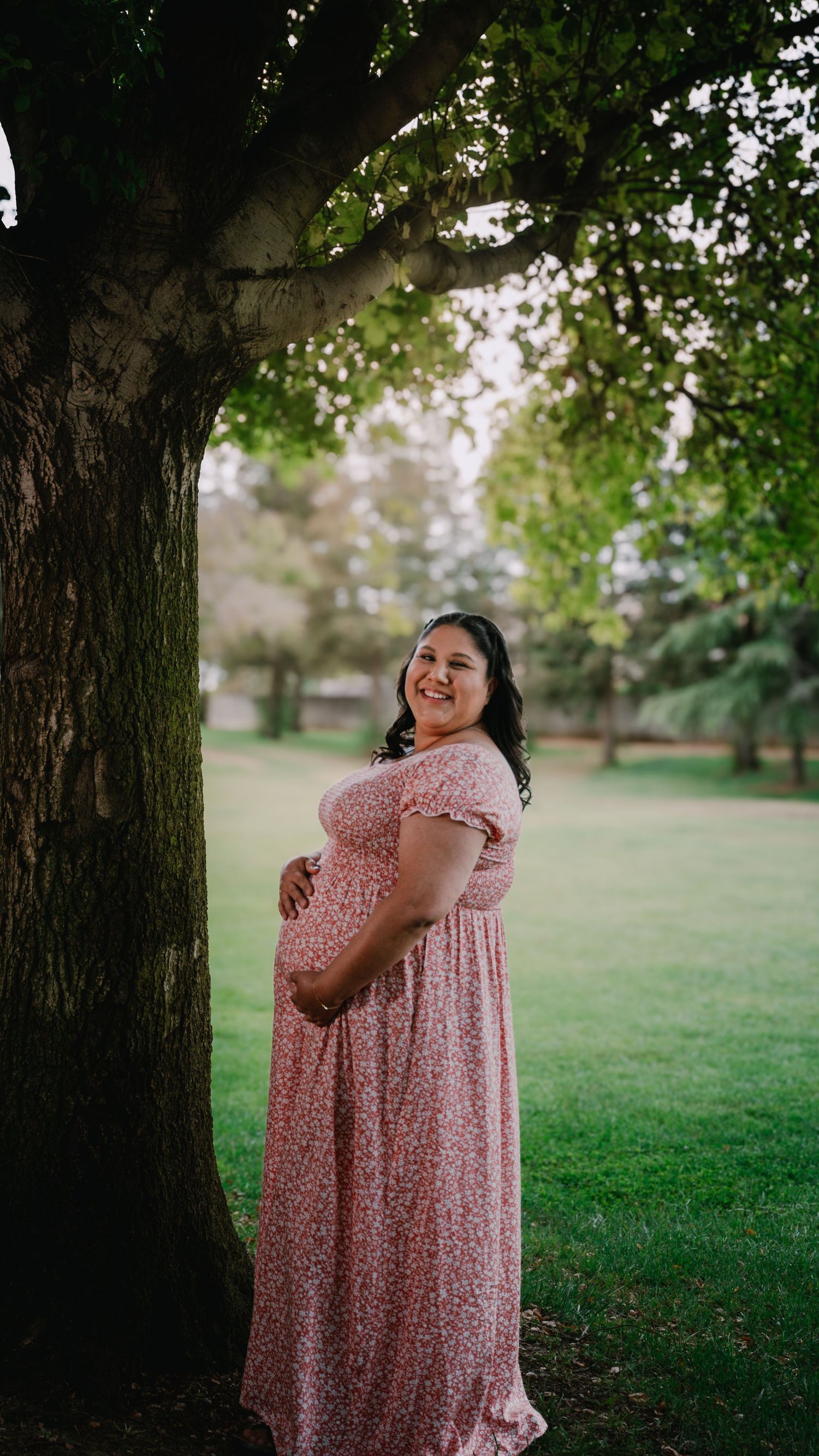 Expectant mother in a pink floral dress poses joyfully under a large tree in a peaceful park setting.
