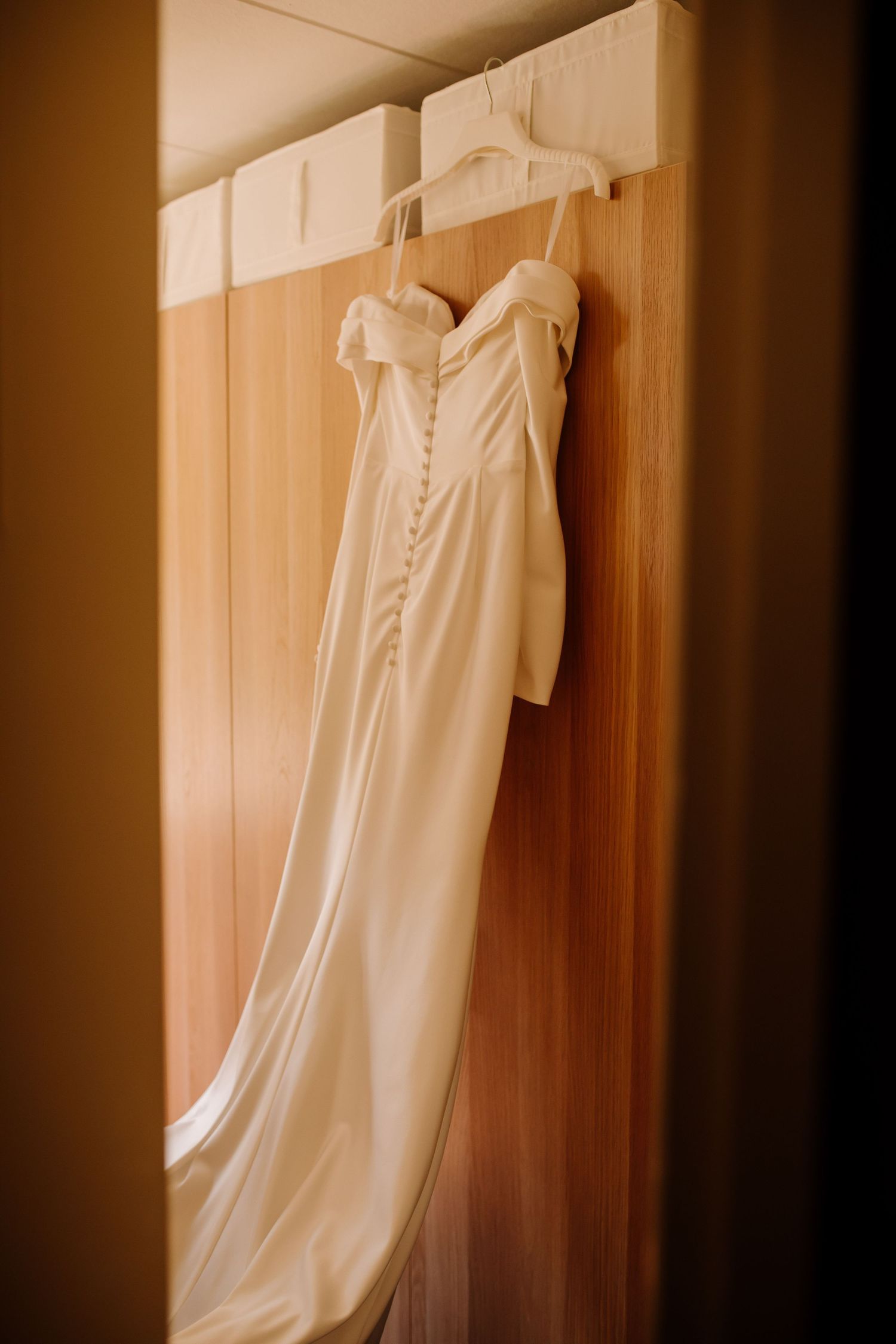 An elegant white wedding dress with buttons hanging on display in a wooden closet.