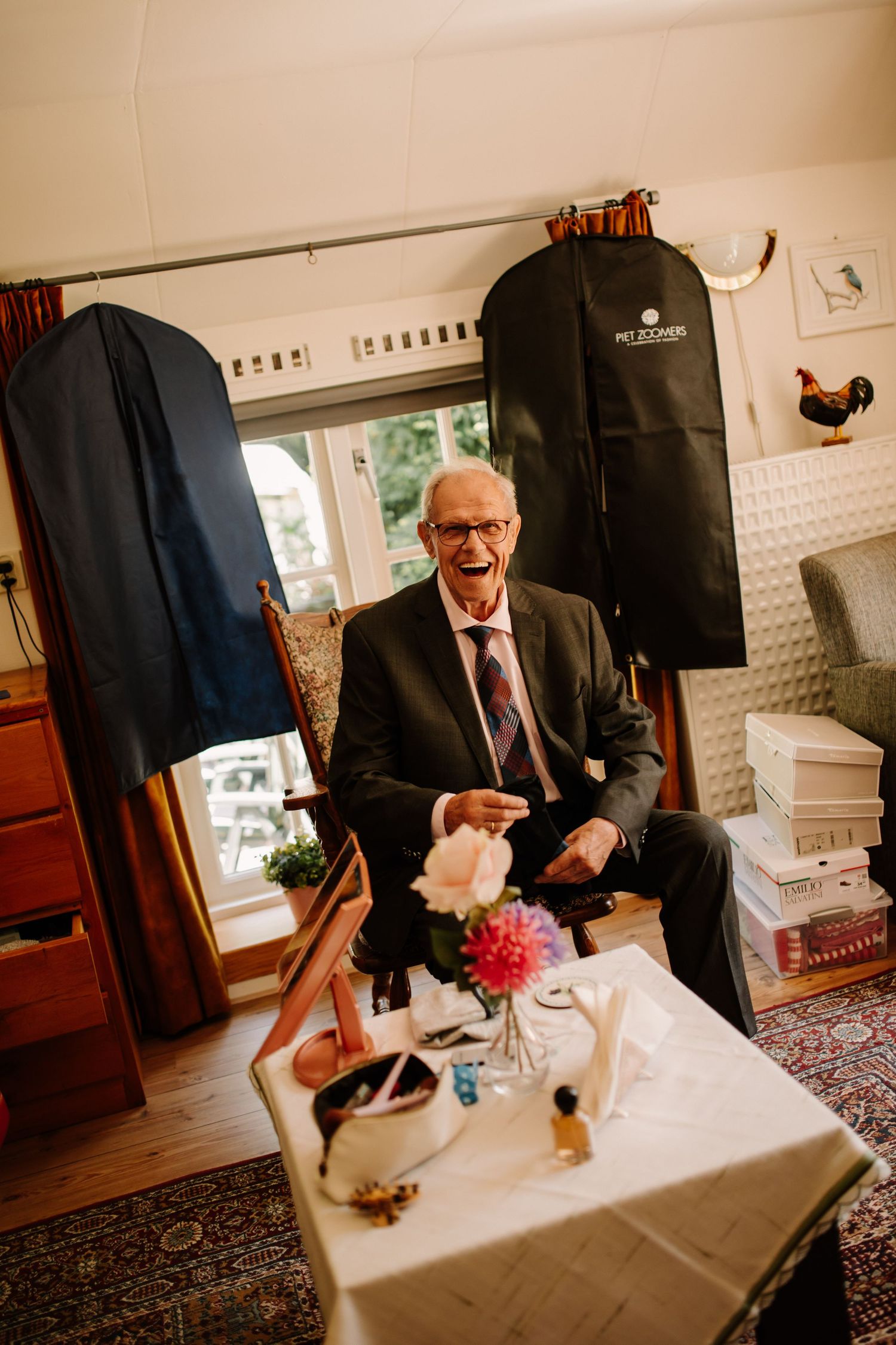 A person seated at a small table with garment bags and boxes in a furnished room.