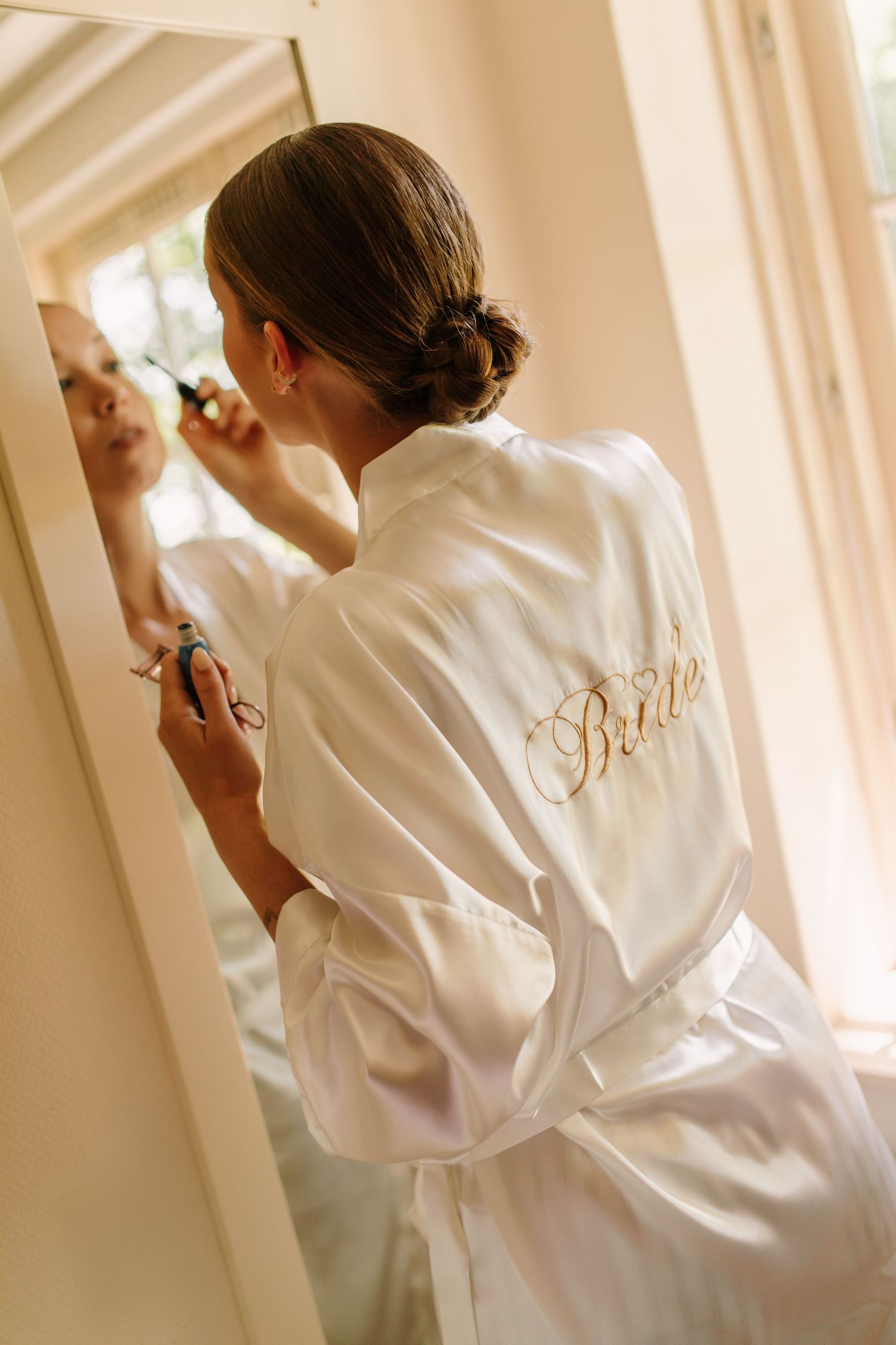 Person in white silk robe with 'Bride' embroidery getting ready in front of a mirror.