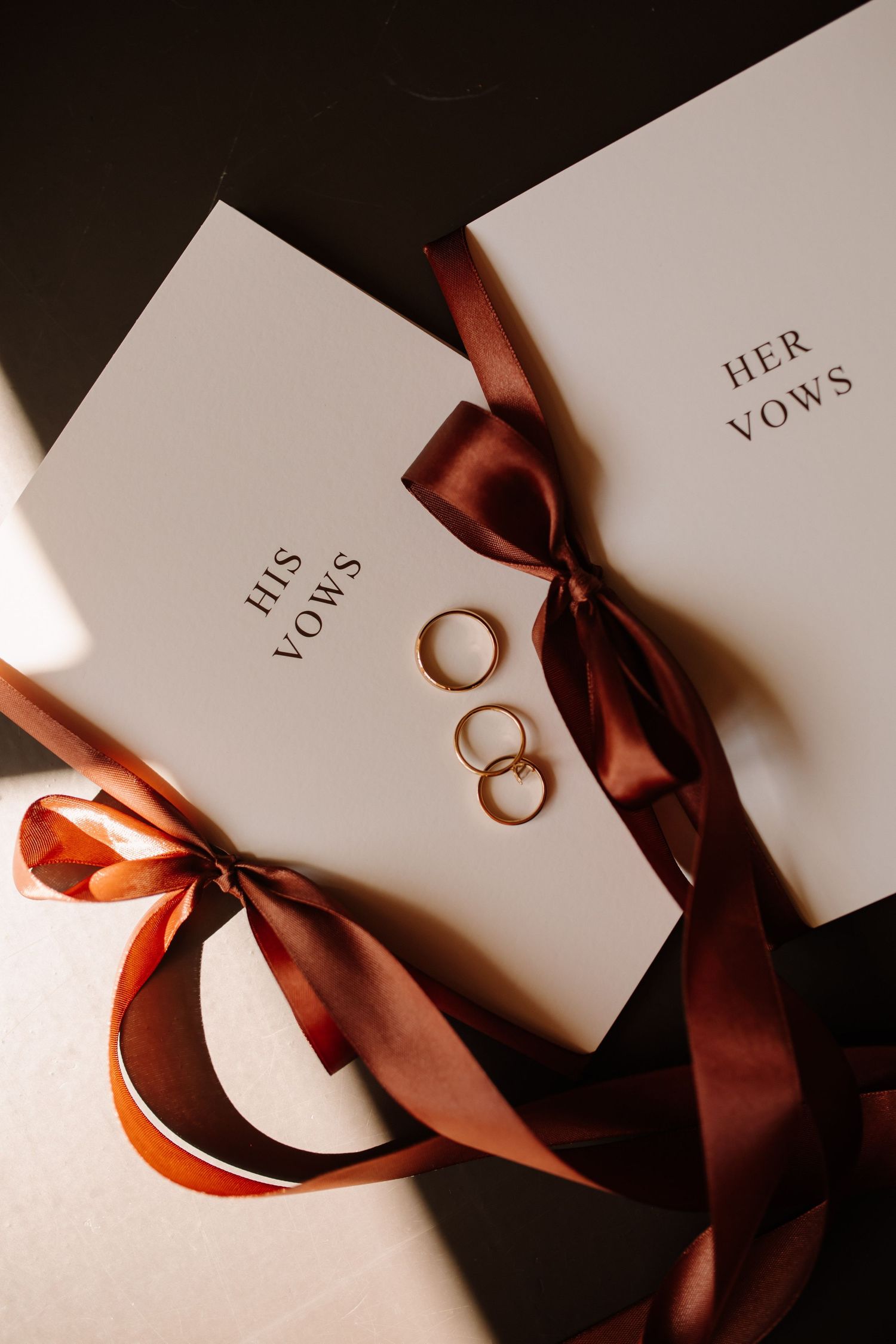 Wedding vow books with burgundy ribbons and gold wedding rings arranged on white surface.