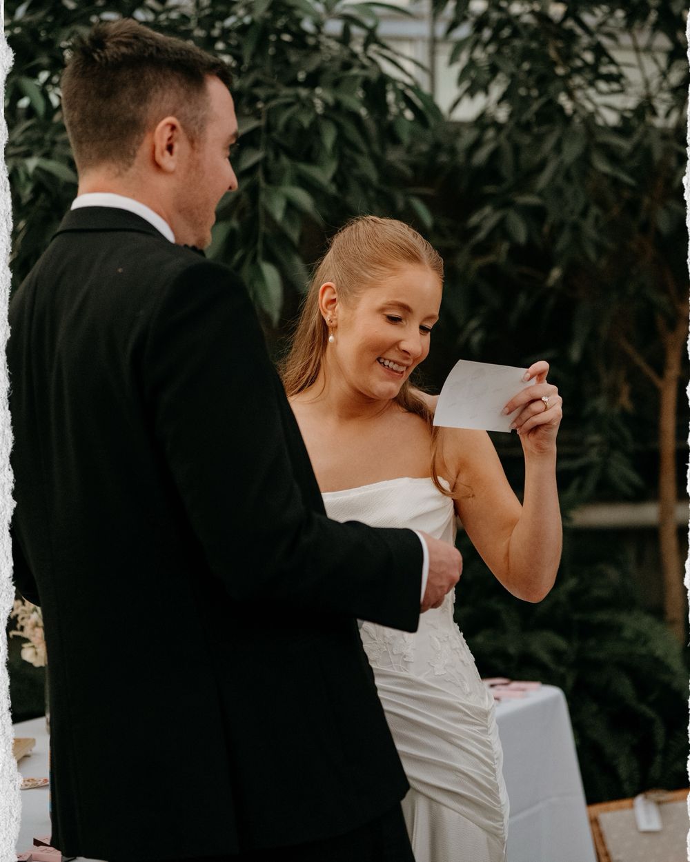Candid moment of a bride and groom sharing laughter during their wedding ceremony against a backdrop of green foliage.