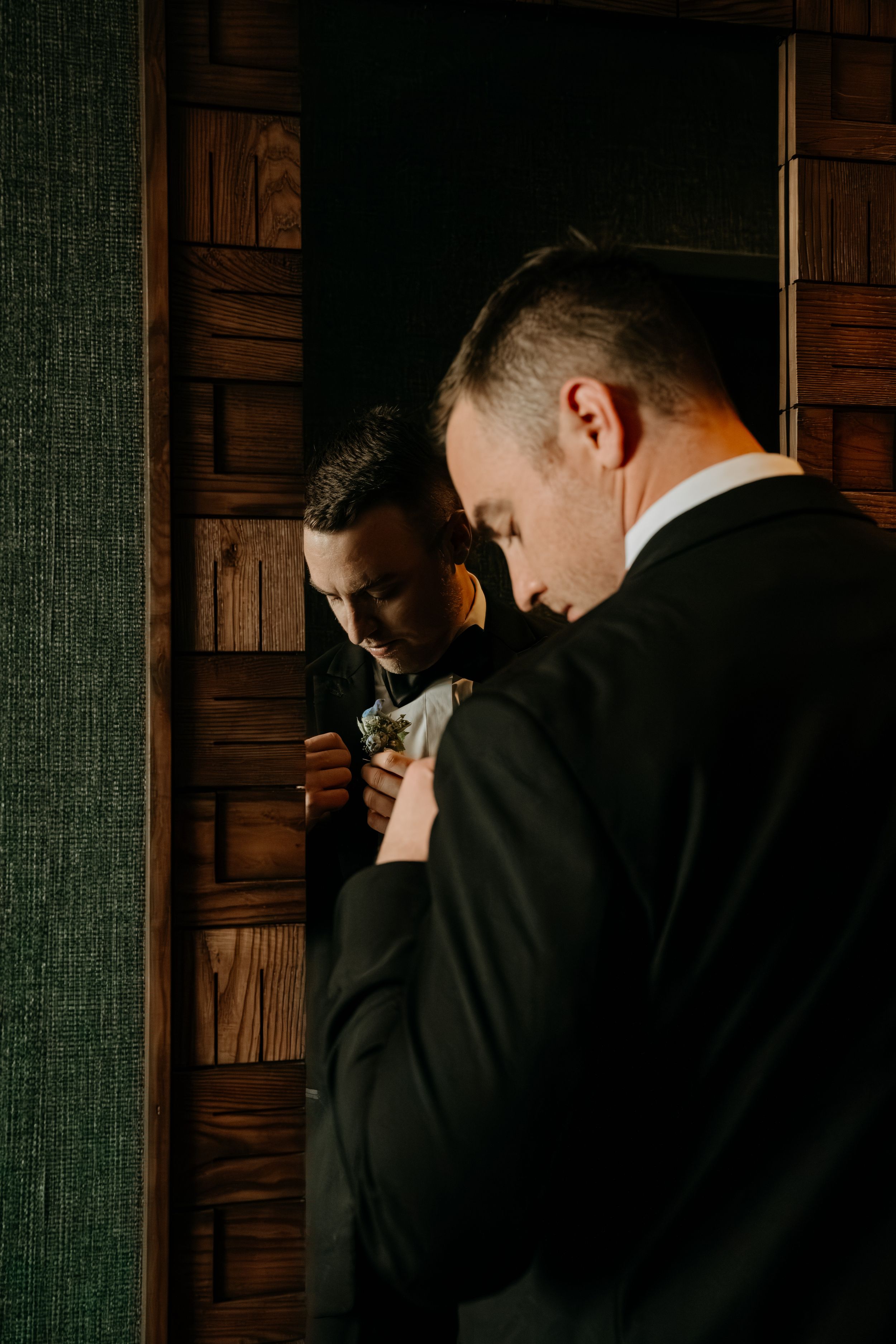A person in a black suit adjusts their tie while looking in a mirror with dark wood paneling behind them.