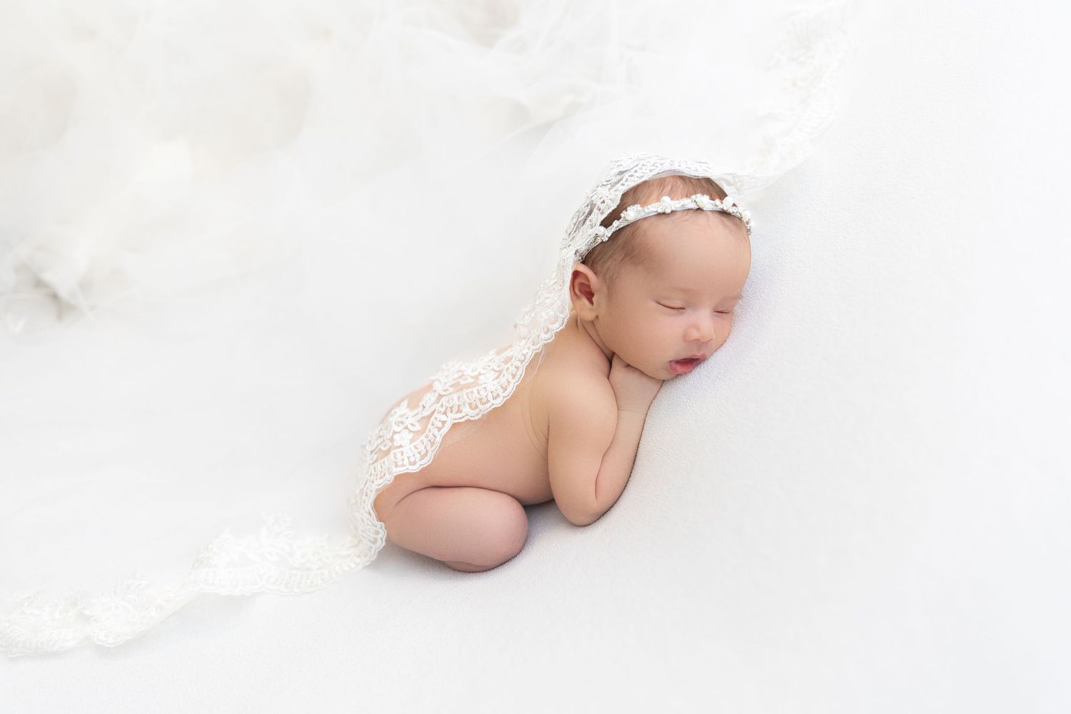 Baby wearing lace accessory lying on soft white fabric background.