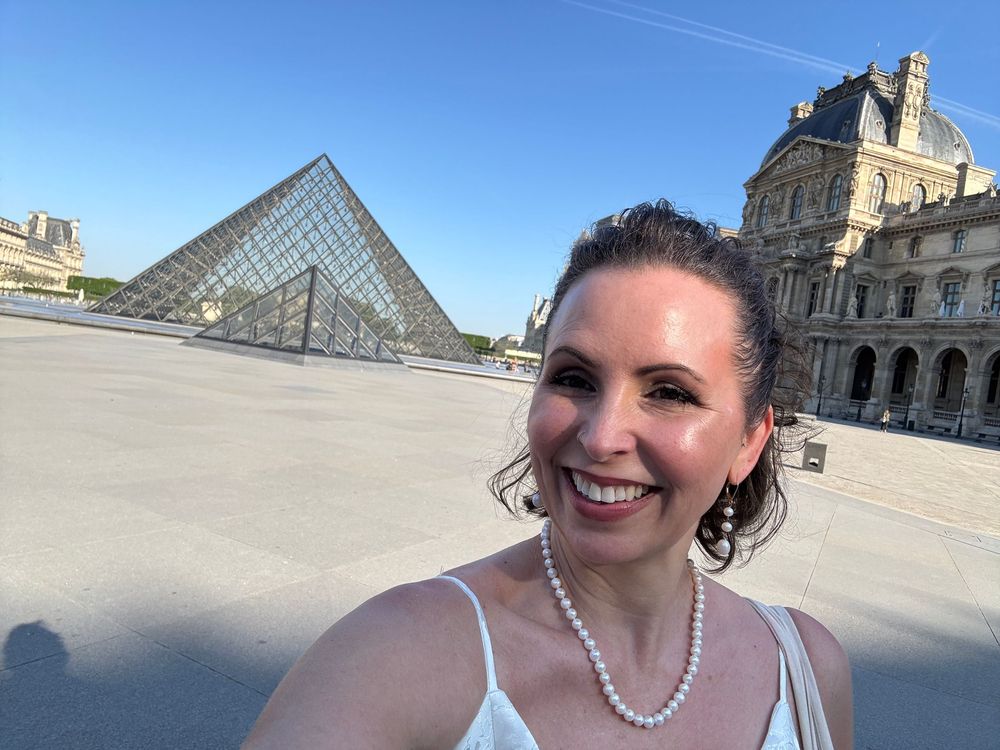 A tourist poses in front of the Louvre Museum pyramid in Paris on a bright sunny day.