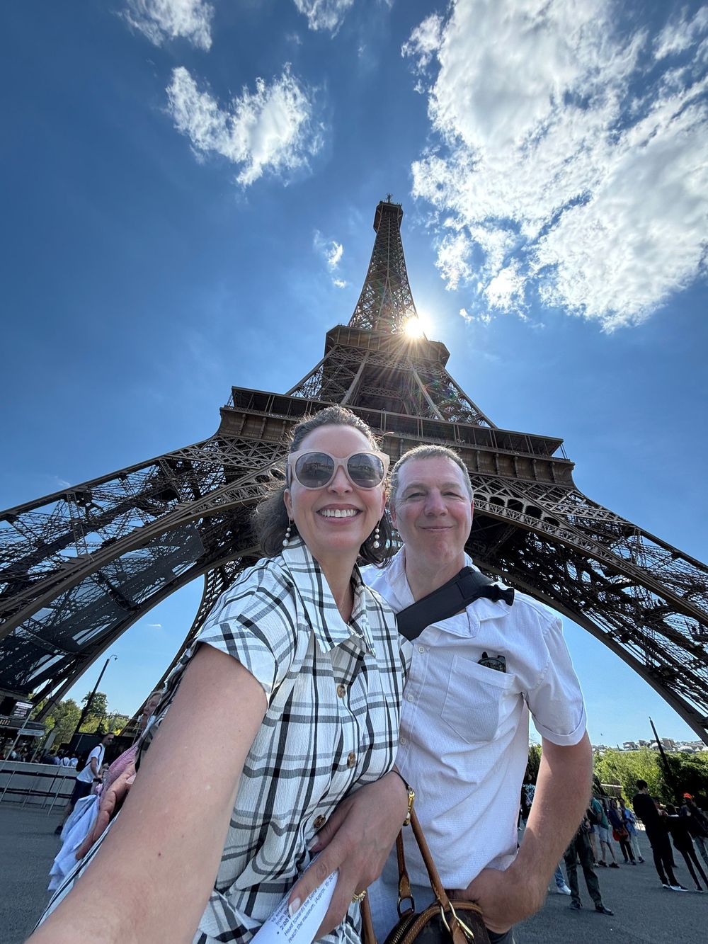 A couple takes a selfie at the base of the Eiffel Tower in Paris on a sunny day with blue skies and white clouds.