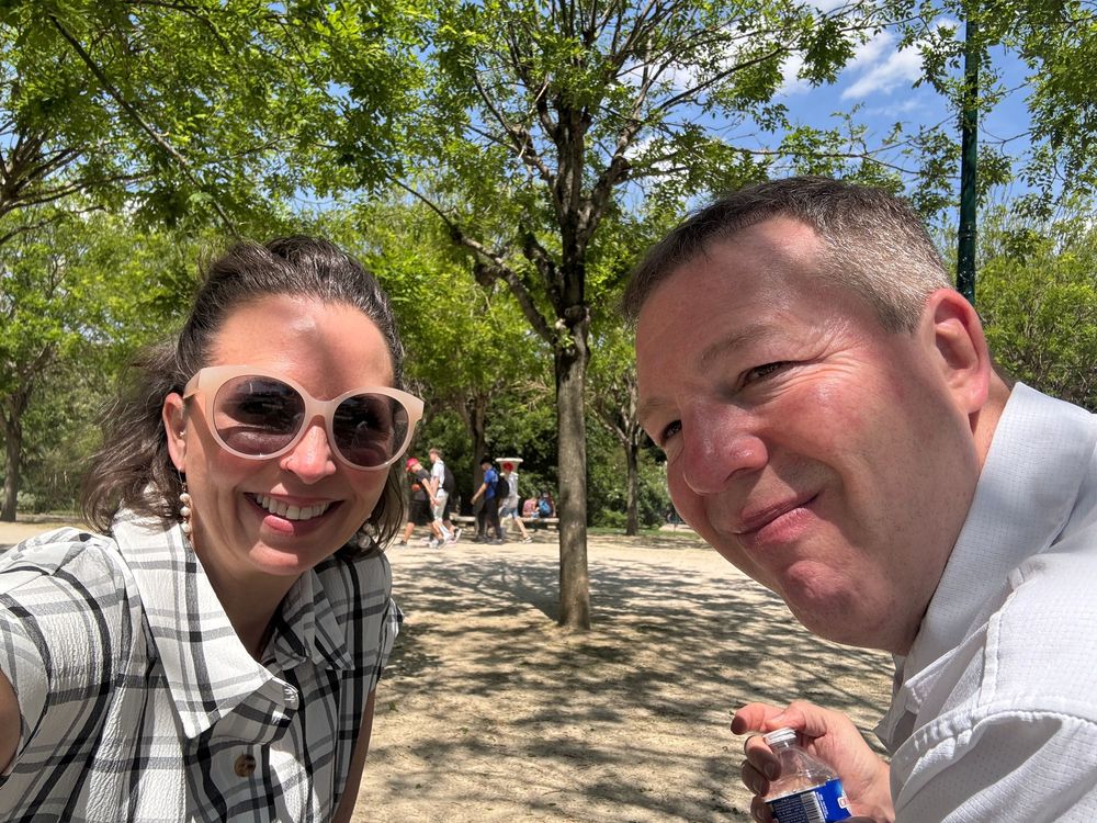 Two people take a selfie together on a sunny day in a park with trees and leaves in the background.