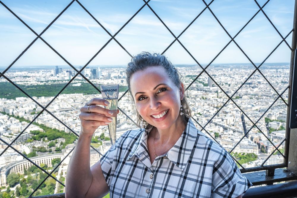 Tourist holding a wine glass and smiling while enjoying panoramic city views from the Eiffel Tower in Paris.