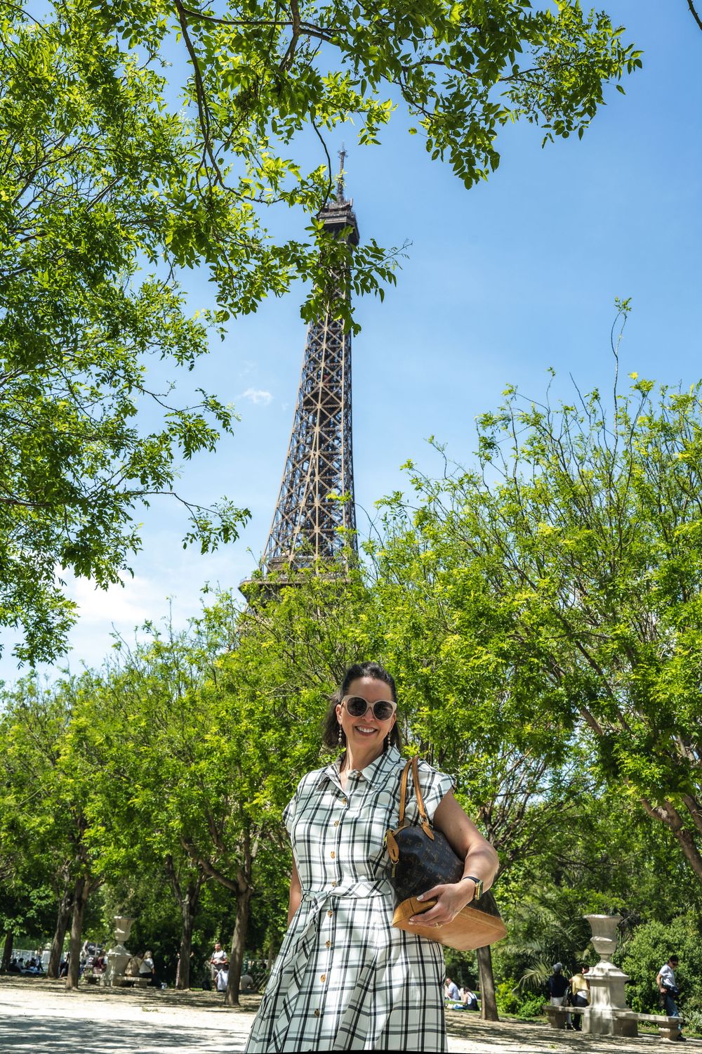 Person in plaid shirt poses near an Eiffel Tower  with green trees in the background at a park.