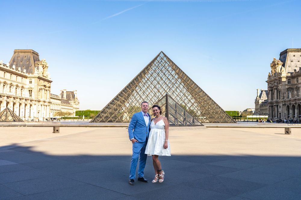 Two people pose in front of the iconic glass pyramid at the Louvre Museum in Paris on a sunny day.