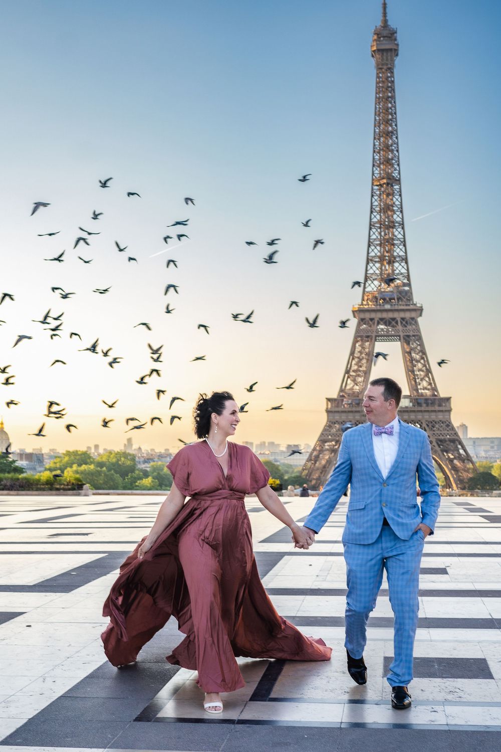 A romantic couple holding hands at Trocadero Square with the Eiffel Tower and flying birds at sunset in Paris, France.