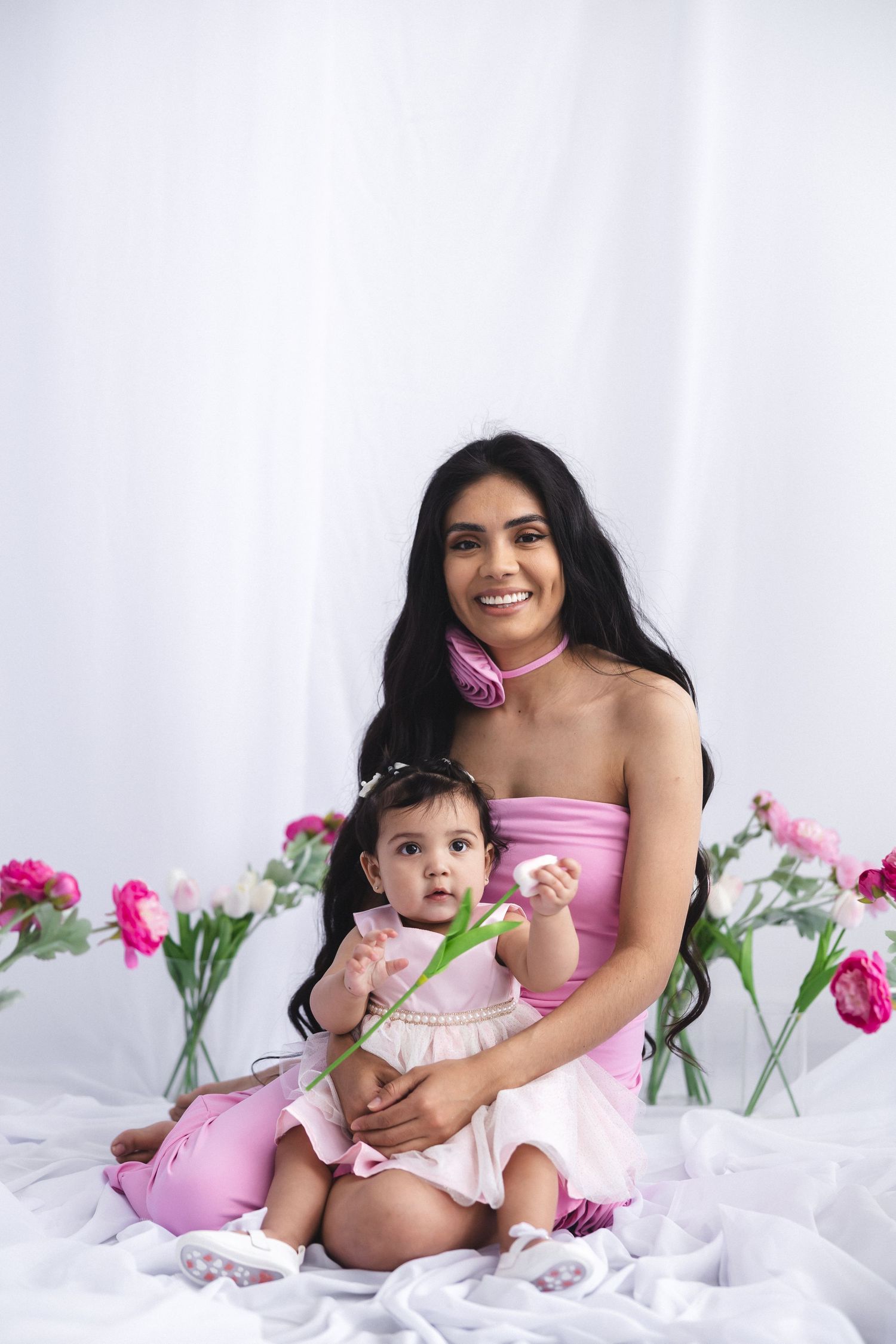 Sweet portrait of mother and baby surrounded by pink flowers on white fabric setting.