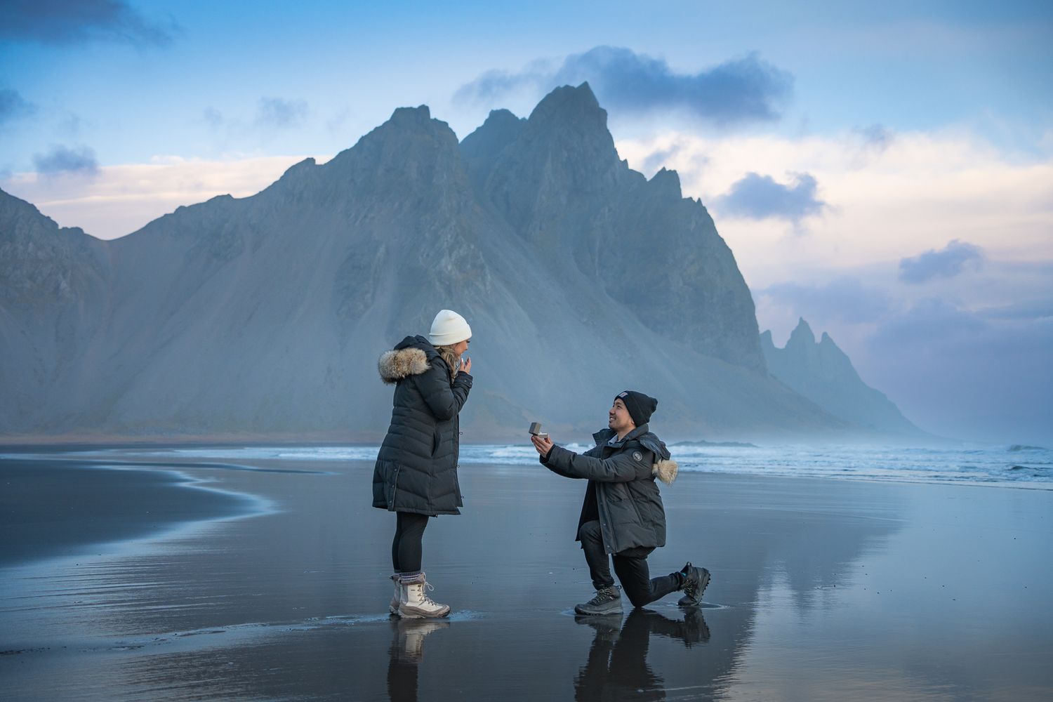 Two people share special moment on black sand beach with jagged mountain peaks in background.