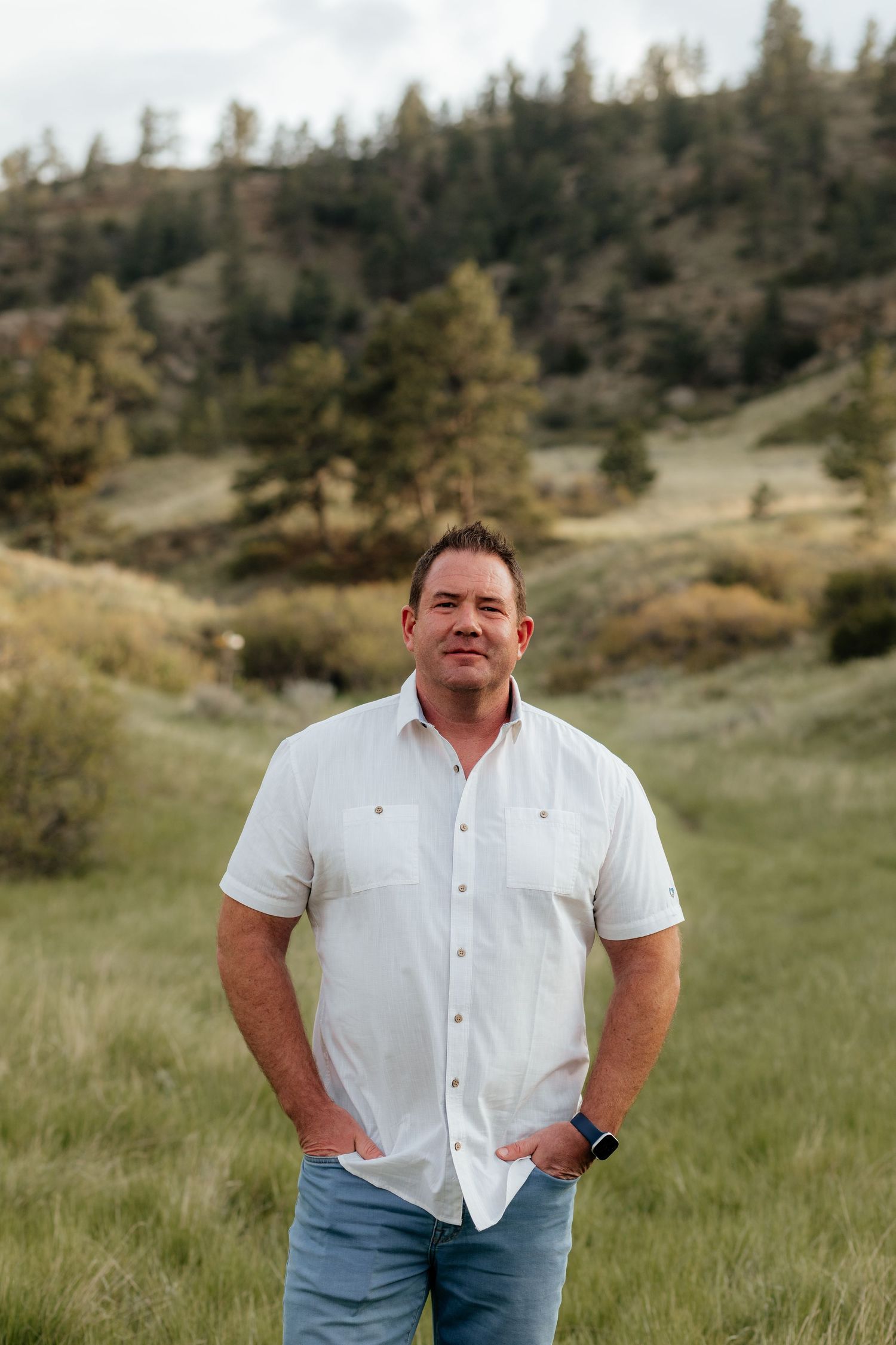 Someone stands casually in a white button-down shirt and jeans against a scenic mountainous backdrop with trees.