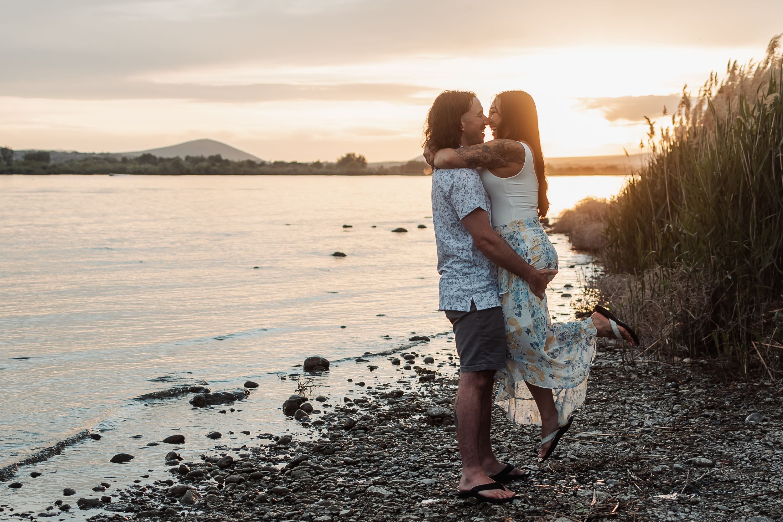 An engaged couple embrace on a rocky shoreline at sunset as golden light reflects off the calm Columbia River water.