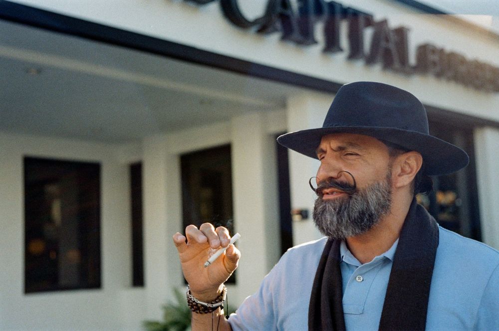 Smiling person wearing a light blue shirt and dark fedora hat stands outside a storefront gesturing with their hand.