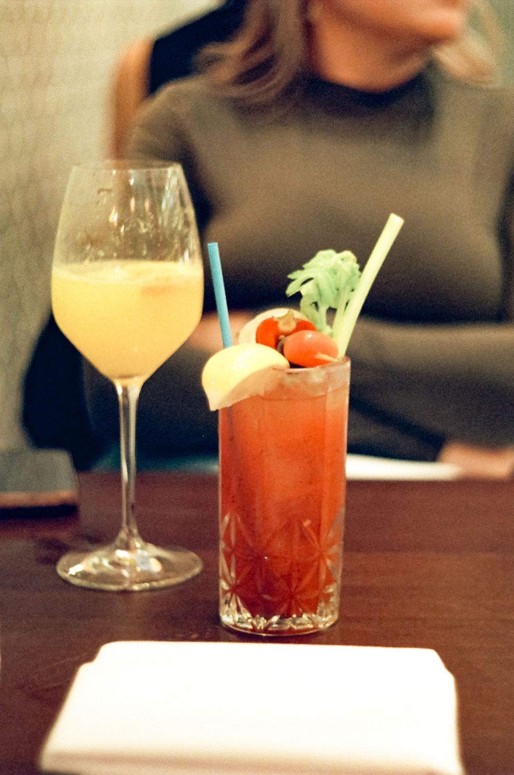 A sequence of four cocktail glasses with varying drinks showing red and yellow hues, garnished with celery and citrus on a bar counter.