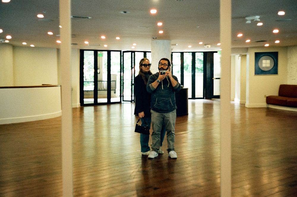 Two people stand in a large empty room with polished wooden floors and glass doors at one end.