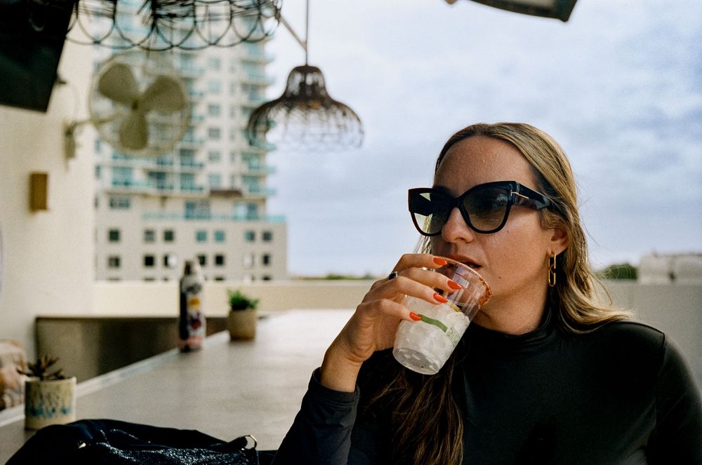 A person in black sweater and sunglasses drinks a beverage on a rooftop terrace with city buildings in the background.