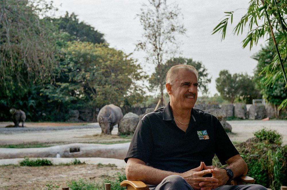 Person sitting outdoors in casual black polo shirt against backdrop of tropical garden and stone structures.