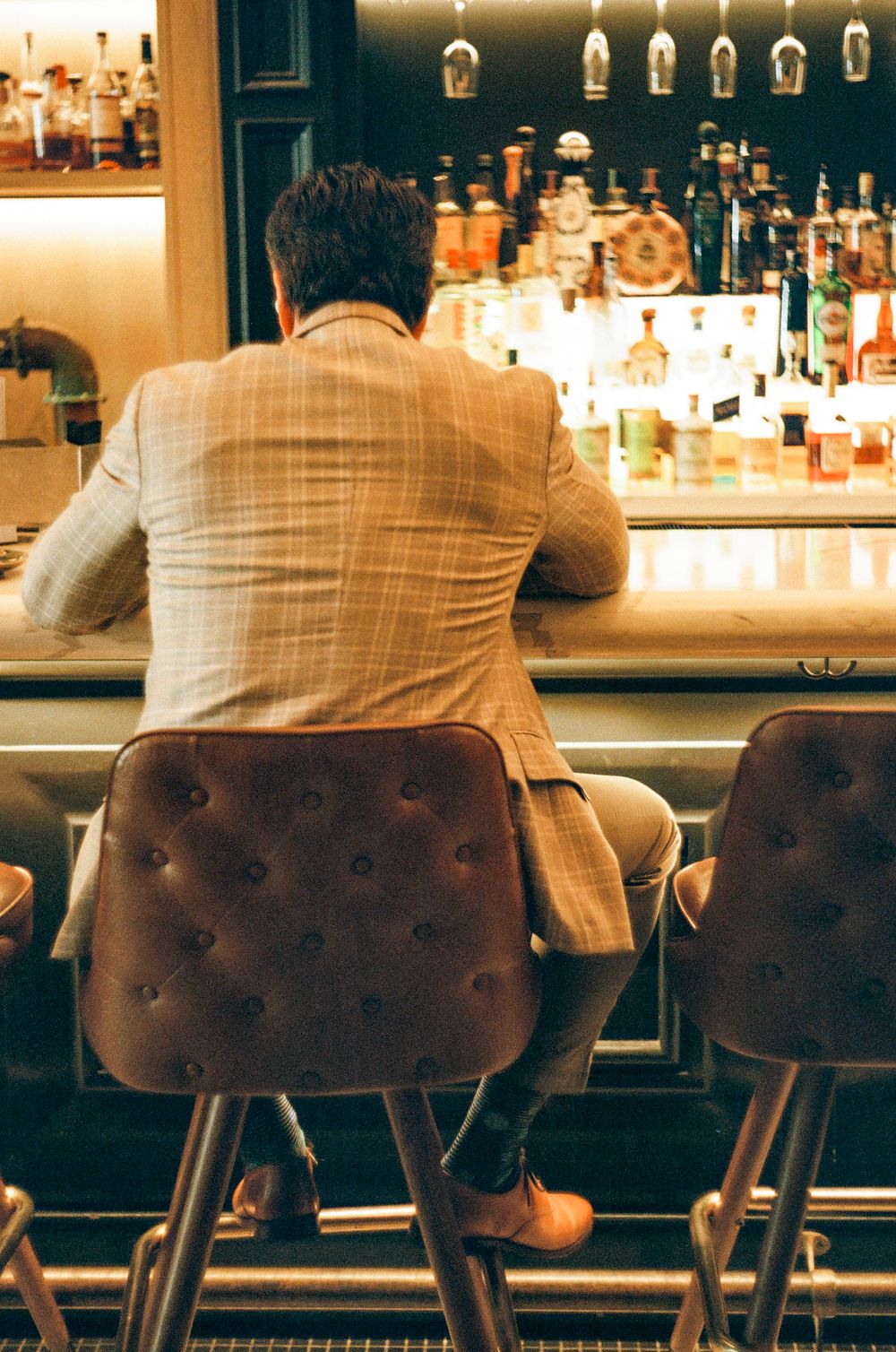 A patron sits at an upscale bar with leather barstools and illuminated liquor bottles in warm ambient lighting.