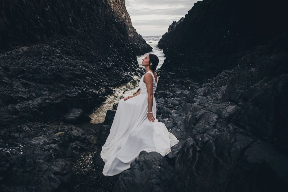A flowing white wedding dress billows in the wind against dramatic rocky cliffs at sunset.