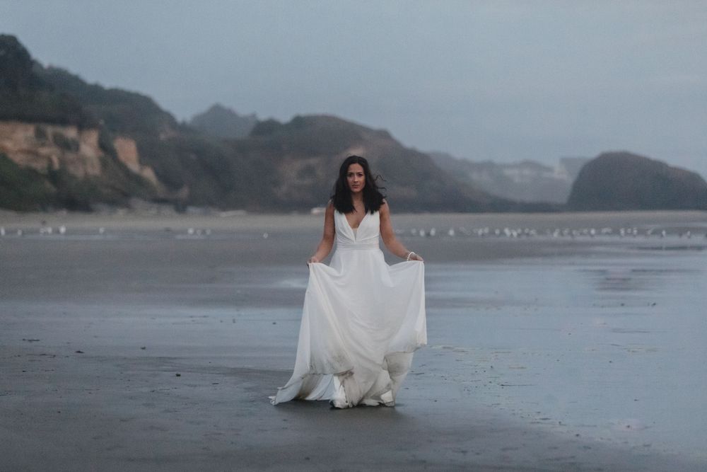 A person in a flowing white wedding dress stands on a misty beach with cliffs in the background at dusk.