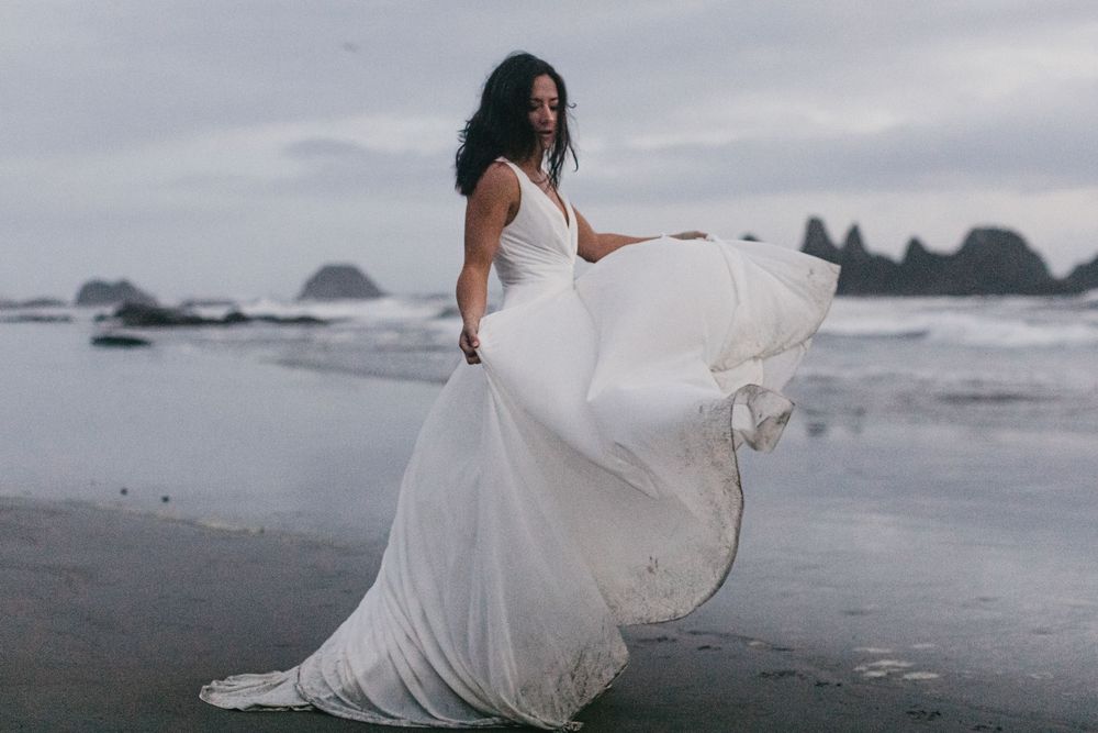 A person in a flowing white wedding dress walks barefoot along a misty rocky beach at sunset.