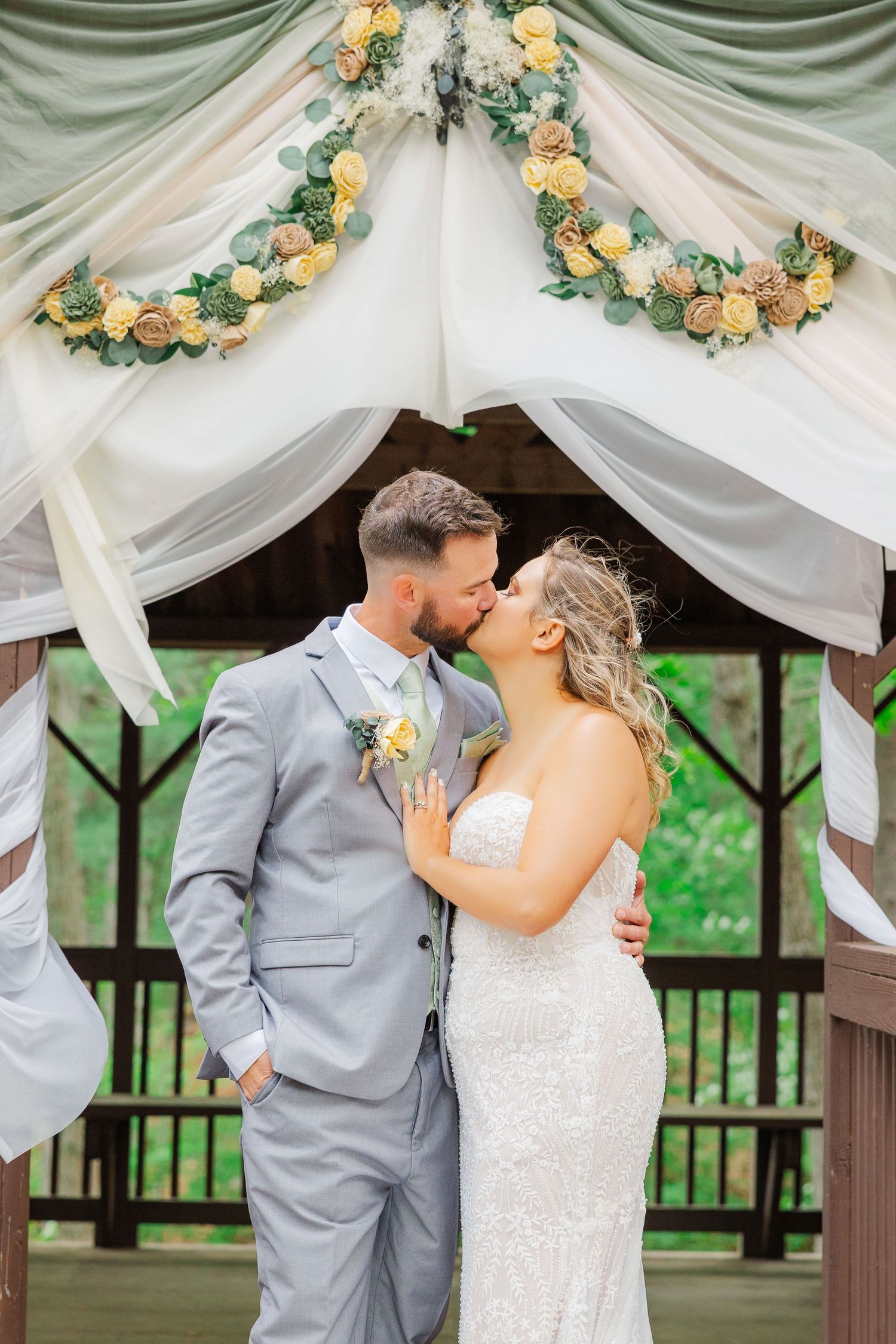 A wedding couple shares a kiss under a white draped gazebo decorated with peach roses and greenery garlands.