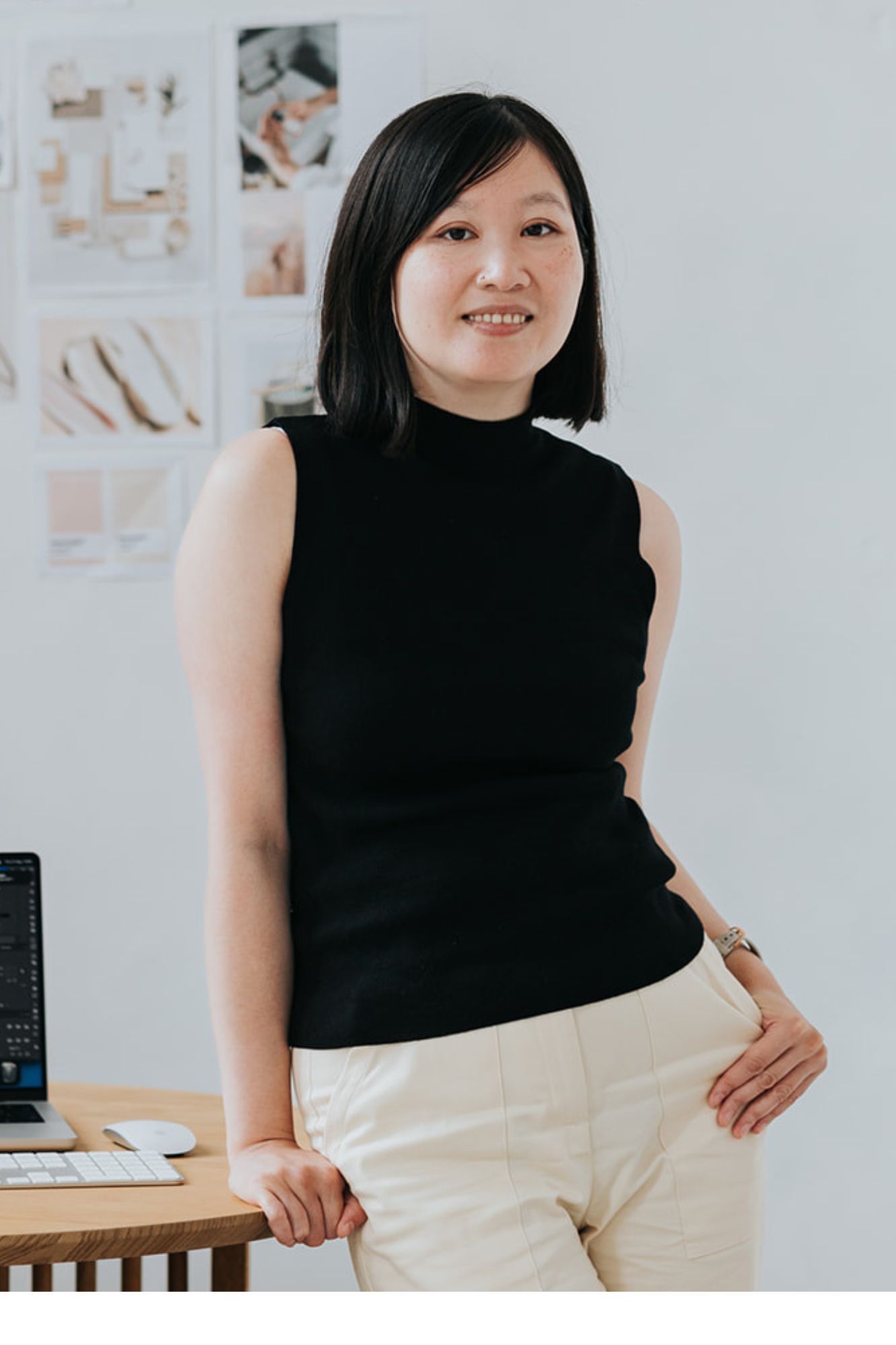 A professional wearing a sleek black sleeveless top and white pants leans casually against a desk in a light modern office setting.